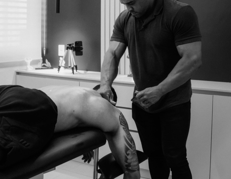 Daniel Torres em p&eacute; no consult&oacute;rio, com as duas m&atilde;os sobre a regi&atilde;o lombar de um paciente curvado sobre a maca. Sobre a foto, um X desenhado a l&aacute;pis marca a panturrilha, onde d&oacute;i, e um c&iacute;rculo marca a lombar, onde o problema come&ccedil;a; um arco liga os dois pontos.