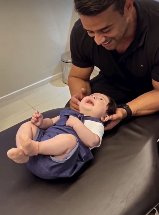 Daniel Torres sorrindo enquanto apoia a cabe&ccedil;a de um beb&ecirc; deitado na maca do consult&oacute;rio.